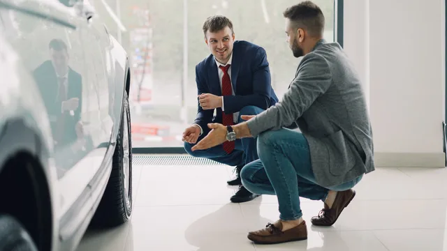 Two men discussing car features in a showroom, kneeling near a vehicle. Measuring readiness before independent customer conversations